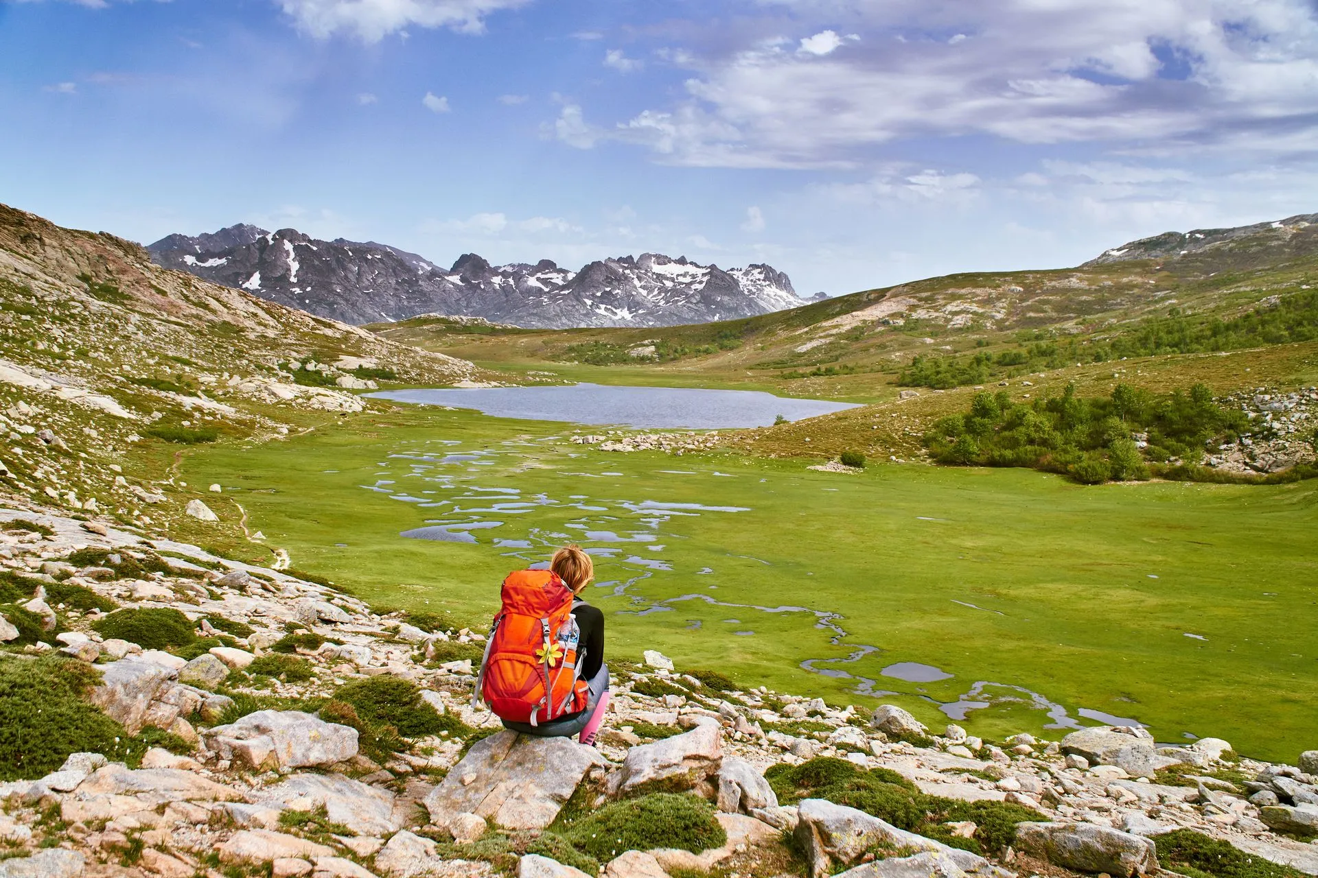 Wanderin mit rotem Rucksack sitzt auf einem Felsen und blickt über eine Hochebene mit Bergsee