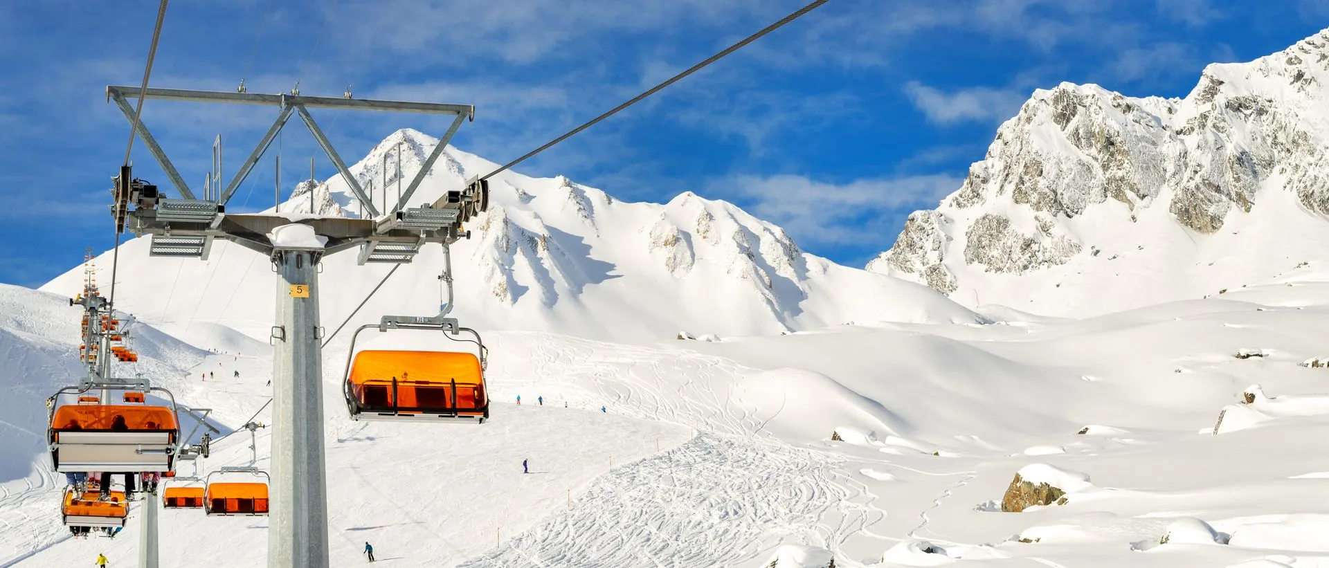 Sessellift mit orangefarbenen Wetterhauben über einer verschneiten Piste vor felsigen Gipfeln